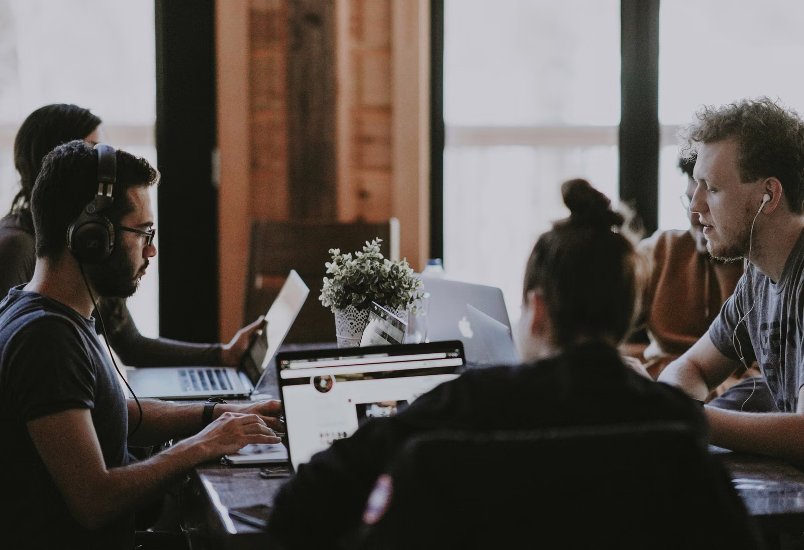 Team members working on laptops around a shared table, one wearing headphones while another talks through a task