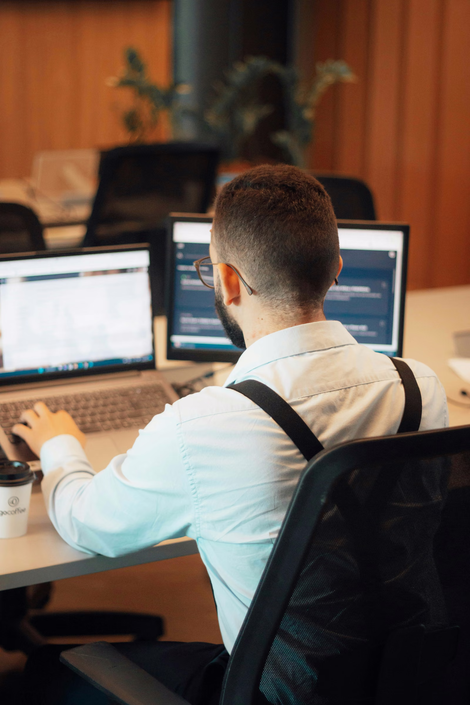 Professional in glasses and suspenders working at a laptop and second monitor in an office